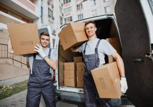 Two young handsome smiling workers wearing uniforms are standing in front of the van full of boxes holding boxes in their hands. The block of flats is in the background. House move, mover service.